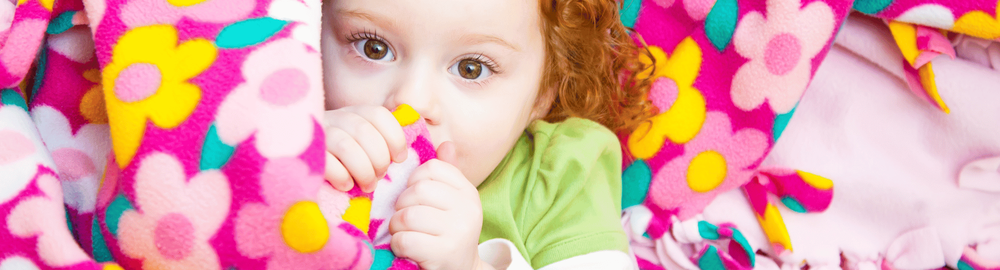 Red headed child with curly hair hiding behind a pink floral tied blanket